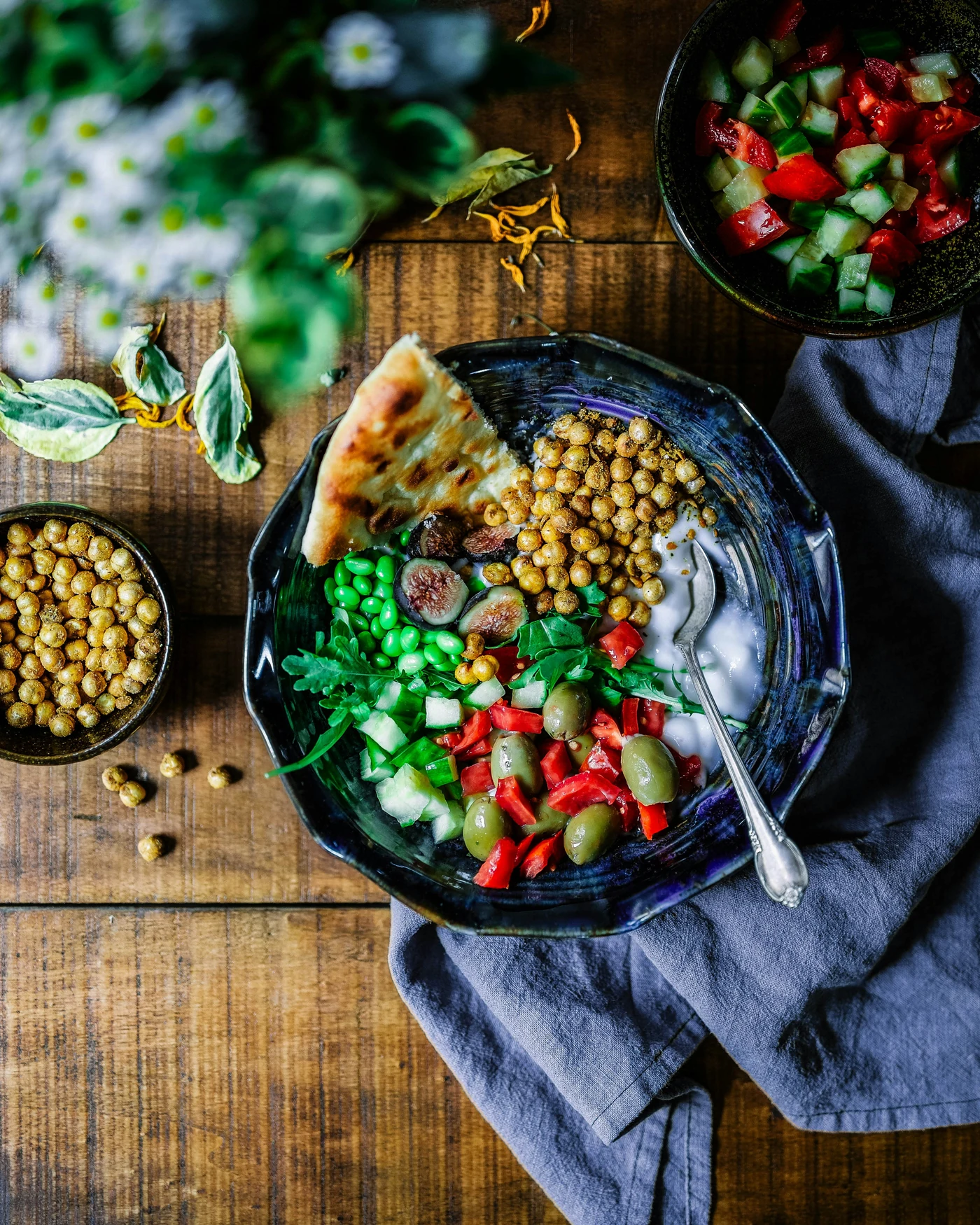 A blue plate filled with chickpeas, greens, colorful vegetables, figs and bread